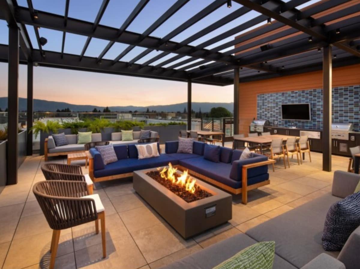 A rooftop terrace features a spacious seating area arranged around a modern fire pit, with flames dancing above the stone base. Comfortable sofas and armchairs provide ample seating, complemented by stylish dining tables in the background. The skyline and mountains create a scenic backdrop.