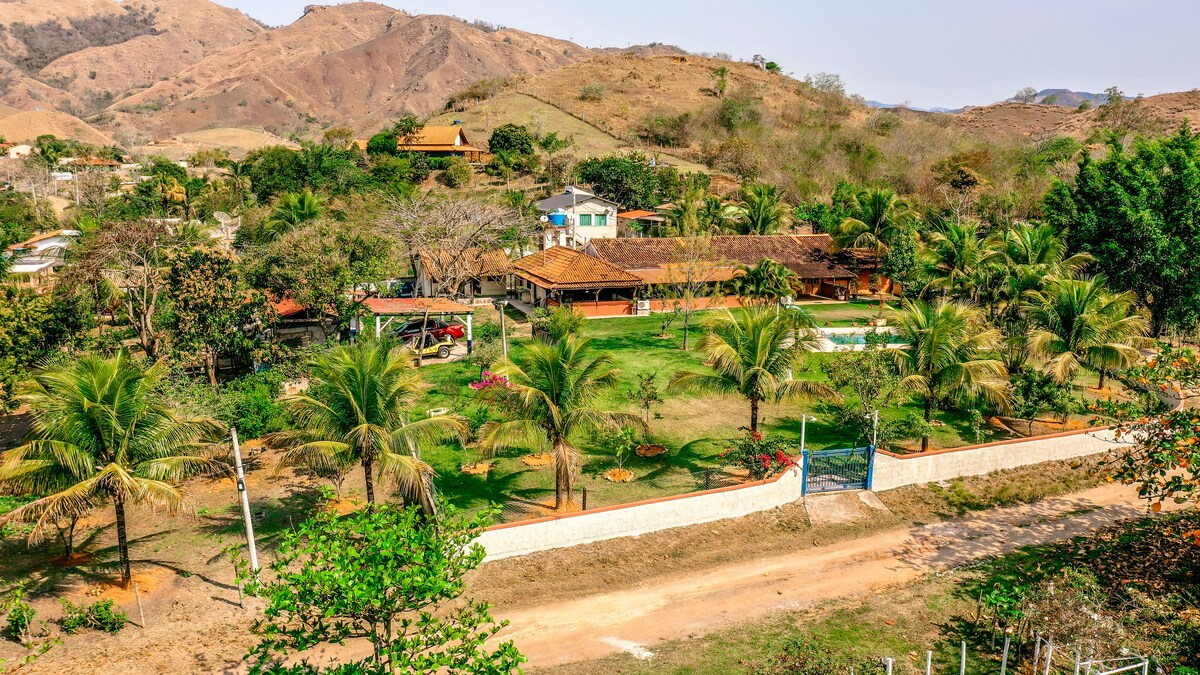 The image showcases a spacious outdoor area surrounded by gently rolling hills. Lush greenery, including palm trees and colorful flower beds, is visible. A well-maintained path leads to the entrance, giving access to the property, which features multiple structures and an inviting pool area.