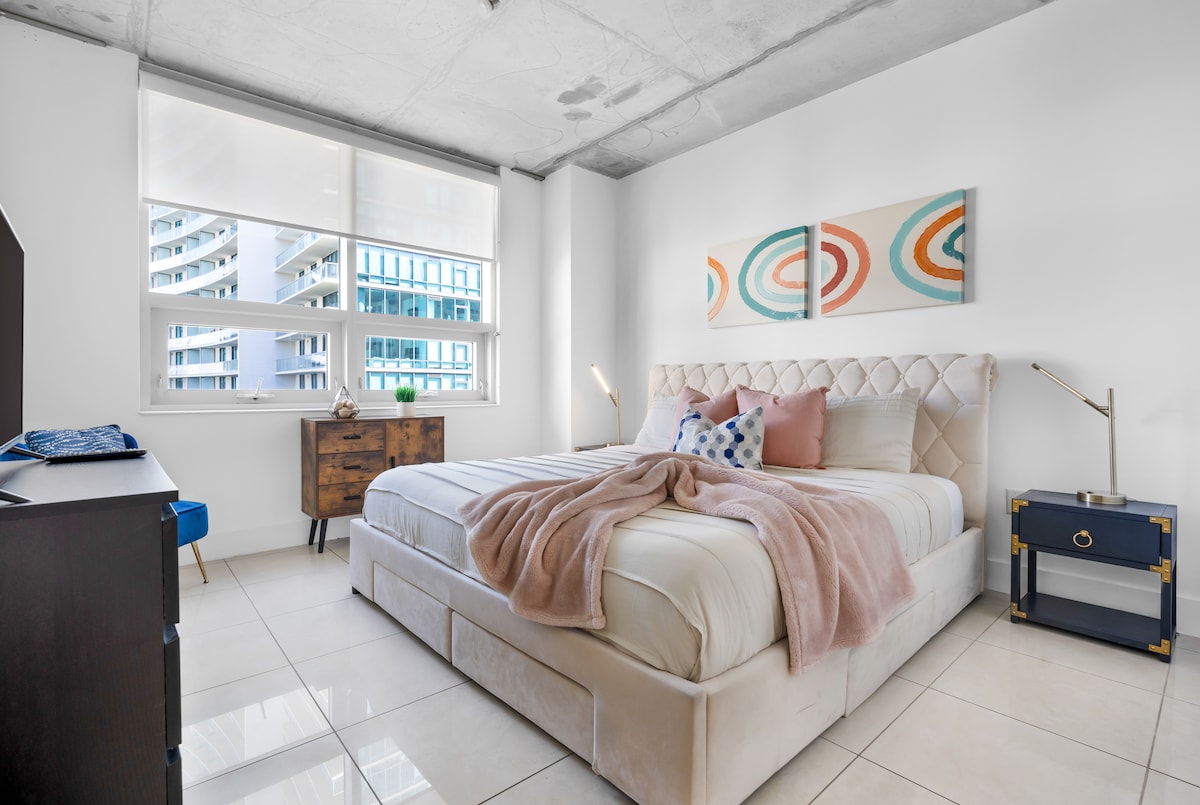 A cozy bedroom features a spacious bed with soft linens and decorative pillows. Two bedside lamps provide warm lighting. Natural light filters through large windows, illuminating the room's neutral palette and modern art pieces on the wall, complemented by simple wooden furniture.