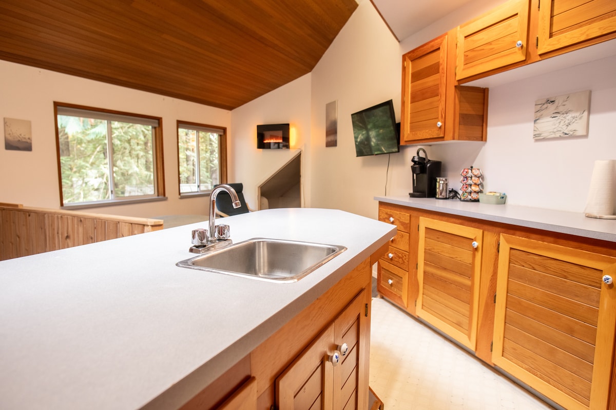 The kitchen area features wooden cabinetry with multiple storage drawers and a countertop with a built-in sink. A coffee maker is positioned nearby on the counter. Windows allow natural light to illuminate the space, while a television is mounted on the wall.