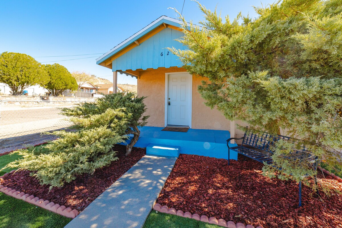 The entrance features a covered porch with a vibrant blue awning. A level walkway leads to the front door, surrounded by neatly landscaped shrubbery and decorative mulch. A metal bench is positioned on the porch, providing a spot to relax outdoors.