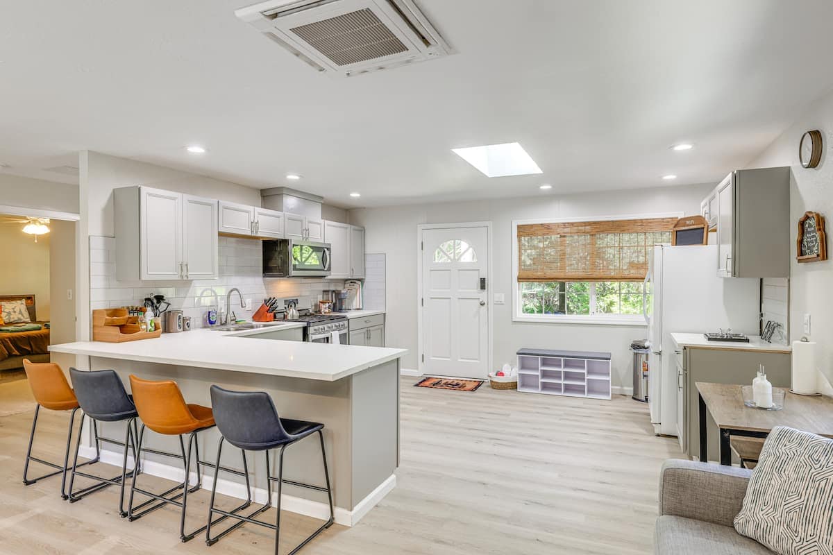 A modern kitchen is visible, featuring a spacious island with three stools, sleek cabinetry, and stainless steel appliances. Natural light floods the area through a skylight and windows, highlighting the welcoming atmosphere of the open layout.