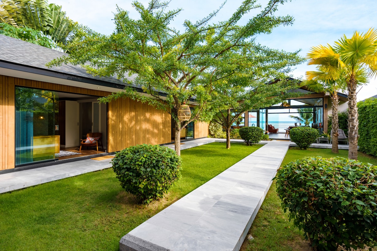 The image showcases a serene outdoor pathway lined with manicured greenery, leading towards a stylish villa. Flanking the path are vibrant trees, including a palm, creating a tranquil ambiance. The sunlit space connects seamlessly with the villa's glass doors, hinting at splendid sea views.
