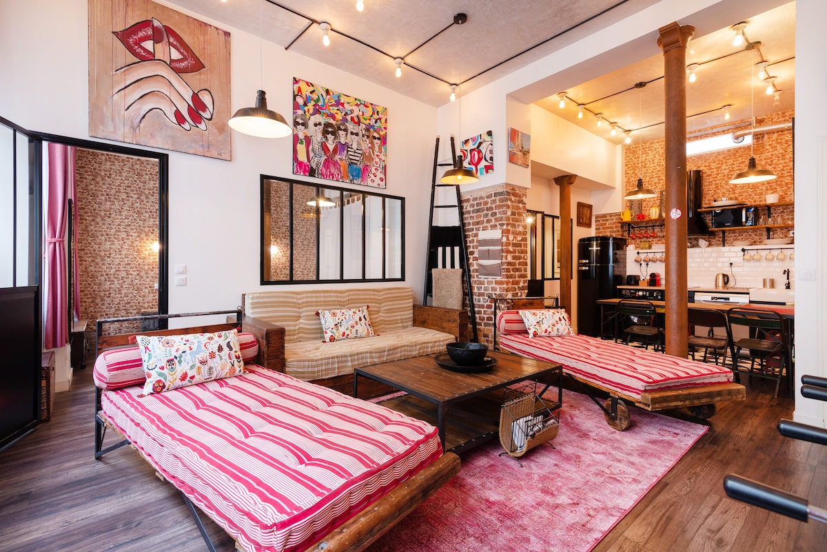 A spacious living area features two striped lounge beds with wooden frames and a cozy area rug. Artistic wall decor enhances the vibrant atmosphere. The open kitchen is visible in the background, complete with modern appliances and a dining space, surrounded by warm lighting.