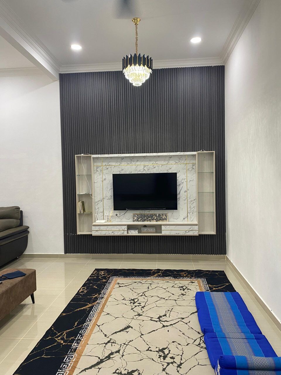 A modern living room is presented, featuring a sleek television mounted on a marble accent wall. An elegant chandelier hangs from the ceiling, while a patterned rug covers the tiled floor. A cozy sofa sits to one side, creating a welcoming environment.