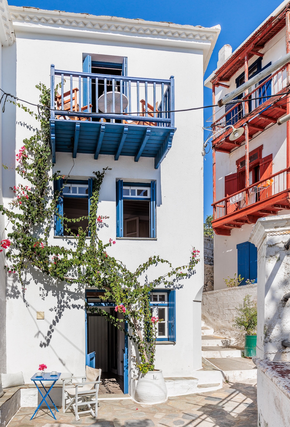 The exterior showcases a traditional three-story house with white walls and vibrant blue balcony railings. Lush green vines climb along the entrance, while a small outdoor seating area is visible. Nearby, a neighboring building features warm red accents, adding to the charming village atmosphere.