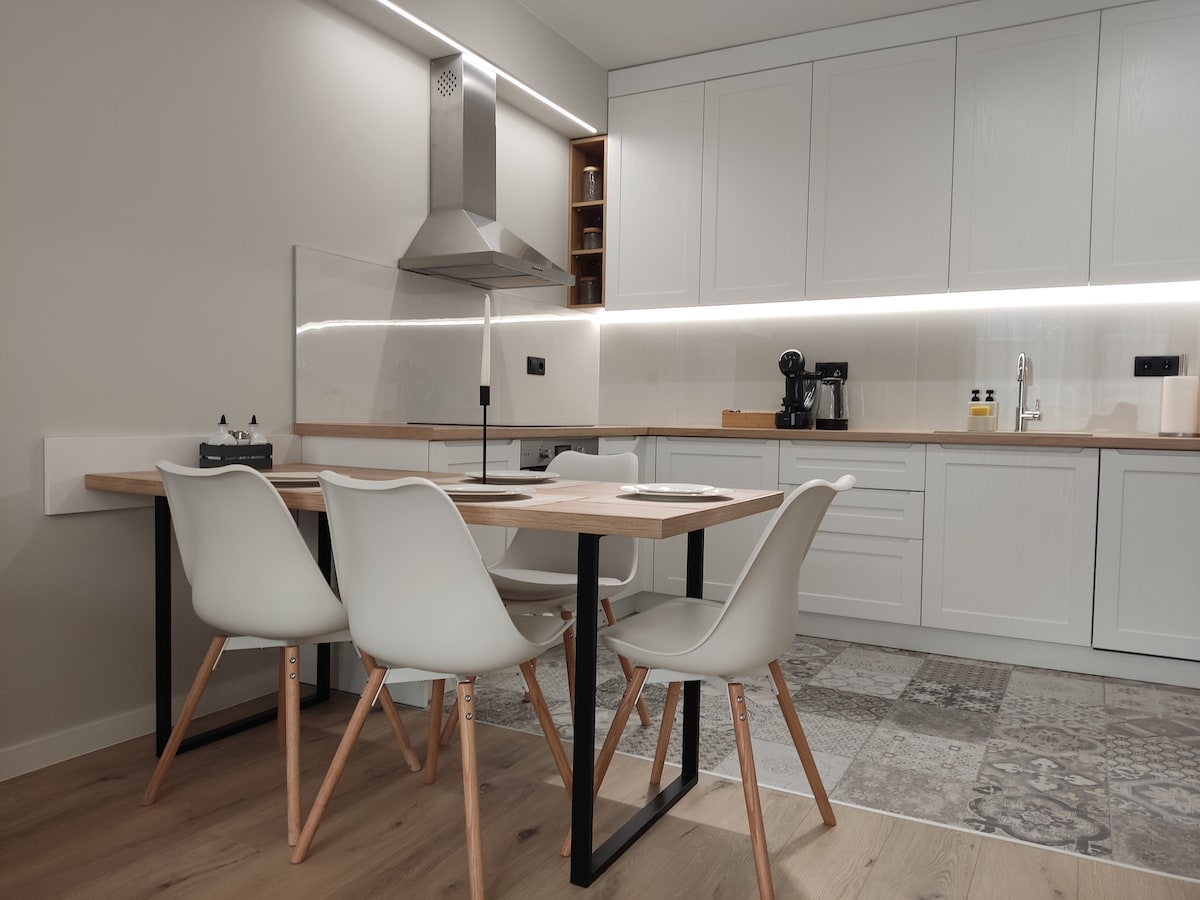 A modern kitchen area features a sleek dining table with four minimalist chairs. The space is illuminated by under-cabinet lighting, highlighting the clean lines of white cabinetry and a stainless steel hood. A coffee maker and other kitchen essentials are visible on the countertop.