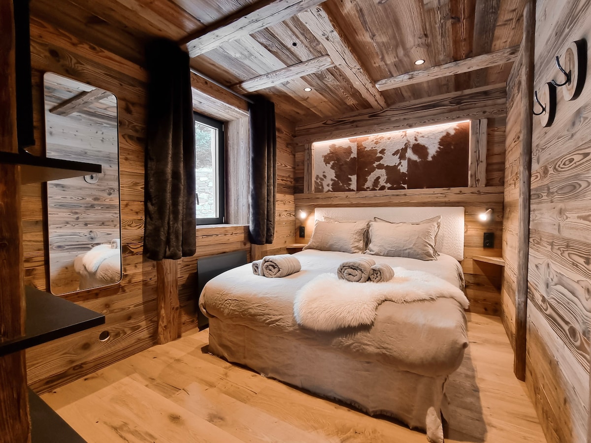 A cozy bedroom features wooden walls and ceiling, creating a warm ambiance. A spacious bed is adorned with soft linens and rolled towels. Natural light enters through a small window, while a full-length mirror and wooden shelves add functional elements to the room.