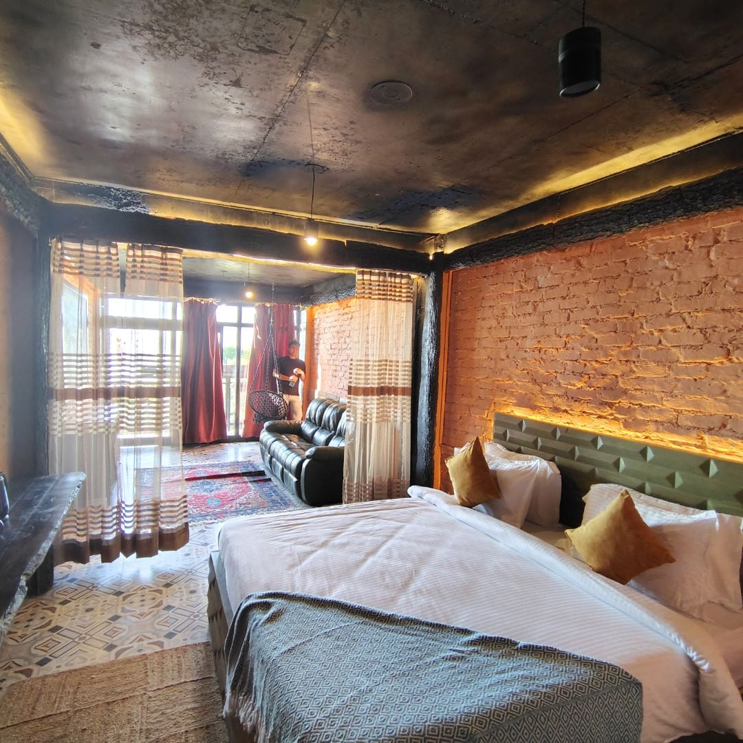 A spacious room features a large bed with plush pillows, complemented by a cozy seating area with a dark leather sofa. Natural light floods in through expansive windows, adorned with sheer and patterned curtains. Exposed brick walls and warm lighting accentuate the inviting atmosphere.