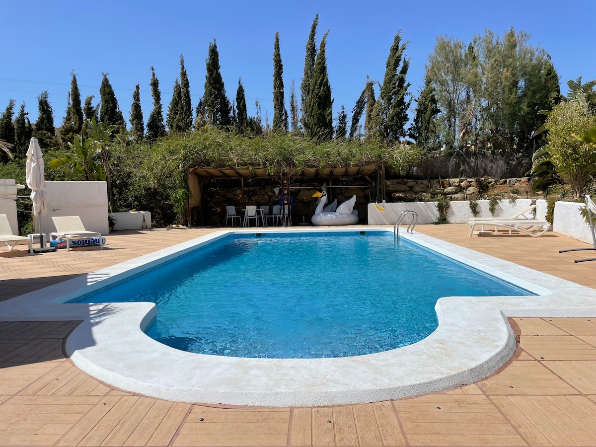 The outdoor swimming pool features a clear blue surface, surrounded by a spacious deck furnished with sun loungers and shaded seating areas. Lush greenery is visible in the background, providing a calming atmosphere. Tall trees stand along the perimeter, enhancing privacy and tranquility.
