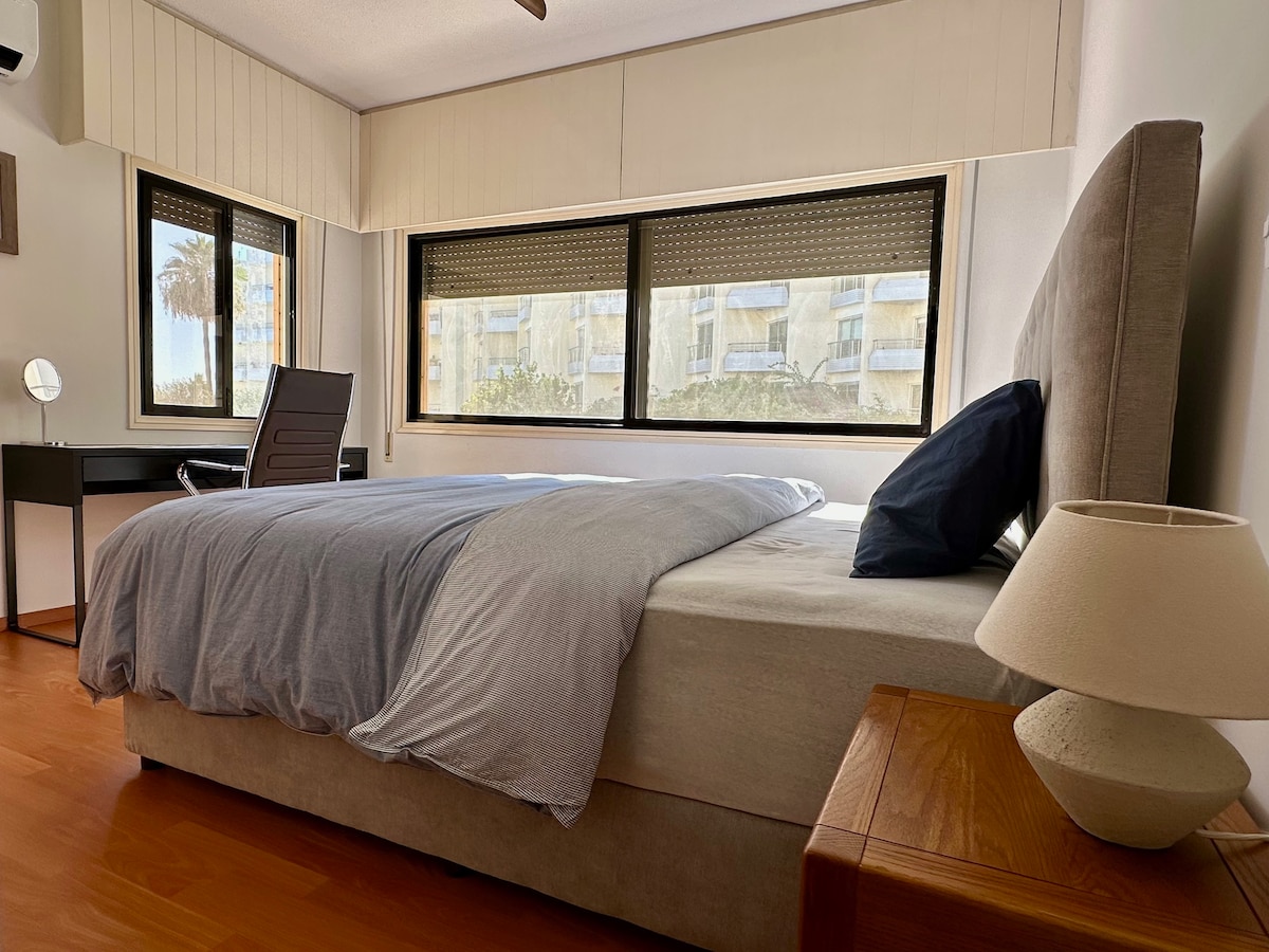 A spacious bedroom is centered around a large bed with a light grey blanket. A small table lamp sits on a bedside table. Natural light floods in through two large windows, showcasing views of palm trees outside. A modern desk with a chair is positioned nearby.