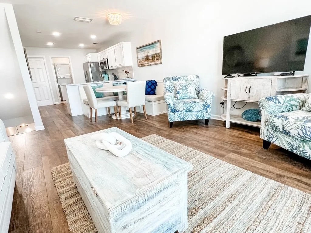 An open living area features comfortable seating with tropical-patterned furniture. A light wooden coffee table sits at the center, complemented by a woven rug. The kitchen area is visible in the background, showcasing modern appliances and a dining table with seating for four.