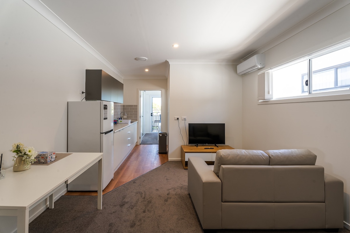 An open living area features a comfortable light-colored sofa facing a flat-screen television. A simple table is present, and a refrigerator with minimal decorations is visible in the kitchen area. Natural light fills the space through a large window.