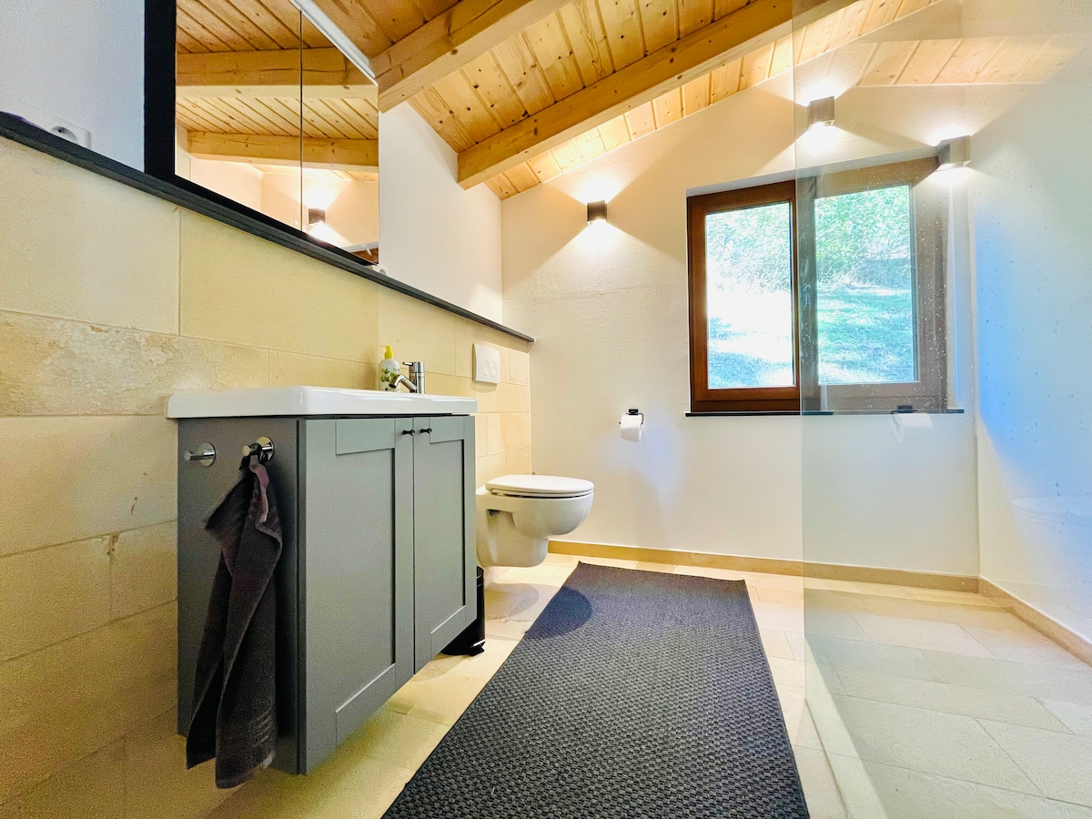The bathroom features a modern vanity with cabinetry and a sink. A window allows natural light to fill the space, while wooden beams add to the ceiling's warmth. A black mat provides comfort underfoot, enhancing the room's thoughtful design.