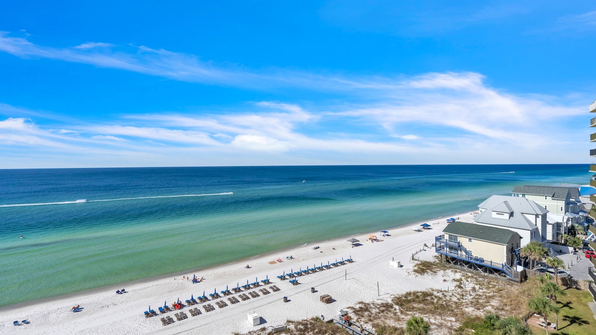The image displays a panoramic view of the beach, featuring soft white sand and clear turquoise waters stretching to the horizon. Beach chairs are arranged near the shore, inviting guests to relax and enjoy the serene coastal landscape under a bright blue sky.