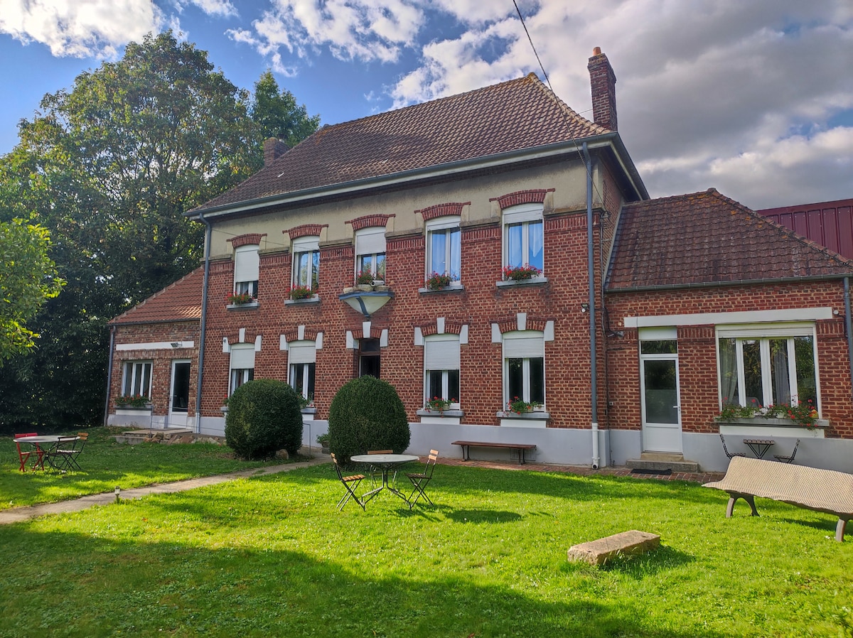The exterior of a charming two-story building is shown, featuring red brick walls and white window frames. Several tables are scattered across the lawn, surrounded by greenery. Flower boxes adorn the windows, adding natural color to the inviting setting.