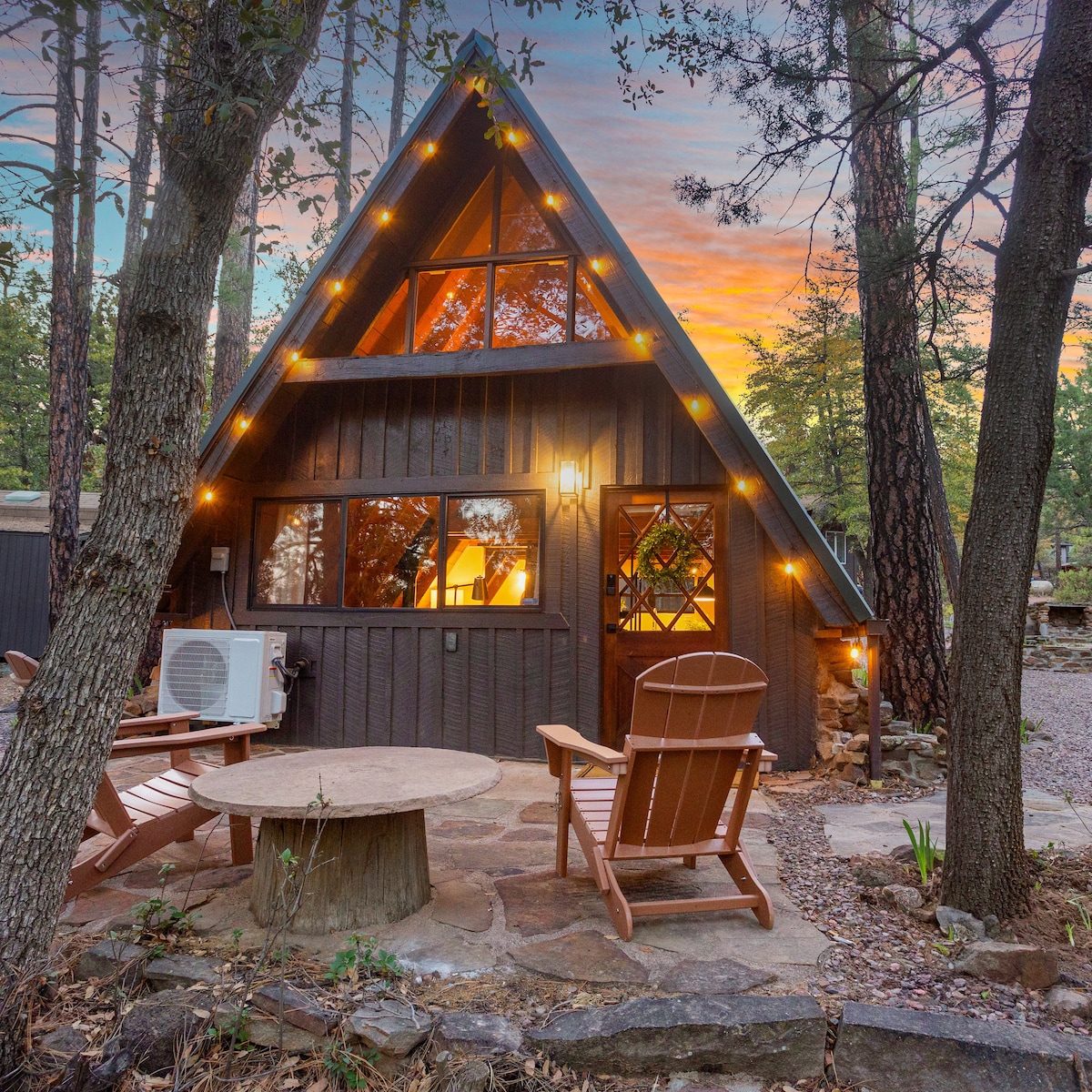 An A-frame cabin is presented with a charming exterior, featuring large windows framed by wooden accents. Cozy seating is arranged on a stone patio, enhancing the inviting outdoor space. Soft exterior lighting adds warmth to the cabin's facade as it stands amidst surrounding trees.
