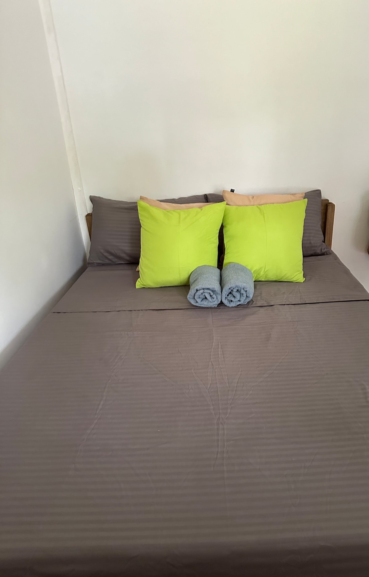 A spacious bed is dressed in soft brown bedding, complemented by two bright green pillows. Rolled towels are neatly placed on the bed, adding a touch of comfort. The light walls create a calm atmosphere, enhancing the room's relaxing environment.