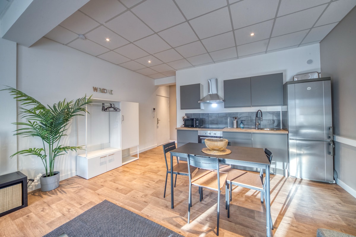 An open kitchen and dining area features a modern grey kitchen with stainless steel appliances and a wooden countertop. A rectangular dining table with black chairs is positioned in the center. A potted plant adds a touch of greenery, and natural light fills the space through overhead lighting.