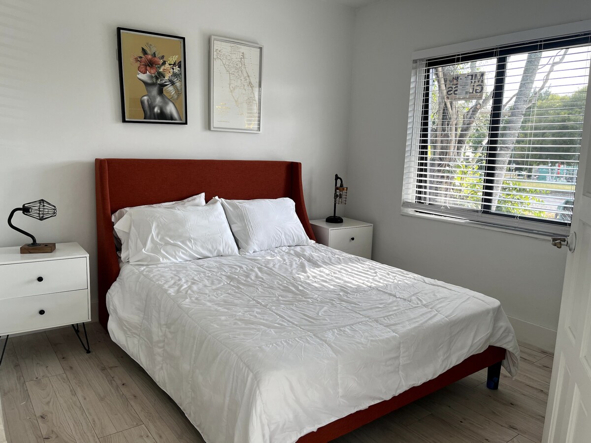 A bright bedroom featuring a comfortable queen bed with white linens. Nightstands are positioned on either side, each with a lamp. Decorative wall art adds detail, and a large window allows natural light to fill the space.