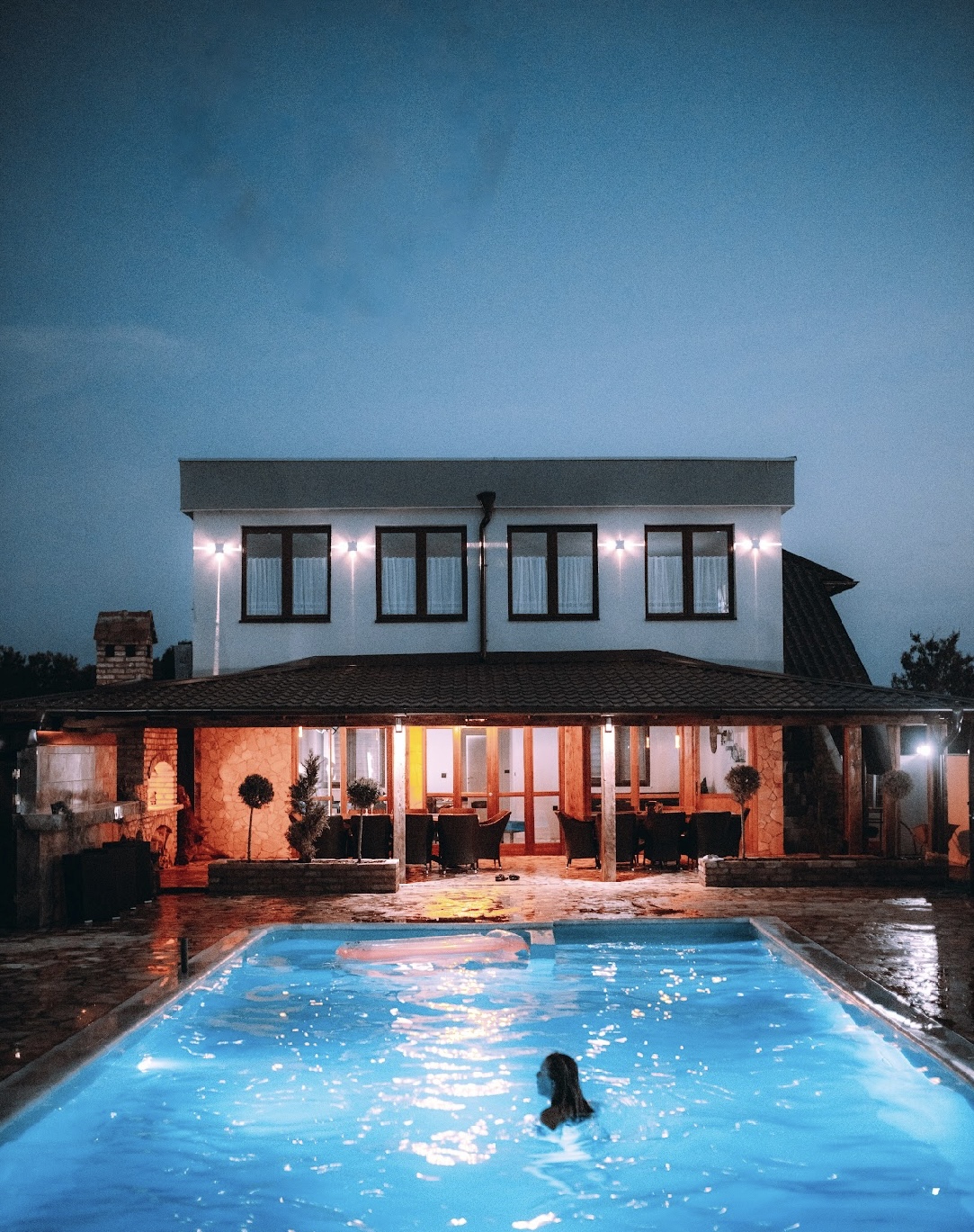 A spacious villa is illuminated at dusk, showcasing large windows and a covered patio area. A tranquil swimming pool reflects warm lighting, while a guest enjoys the refreshing water. The outdoor space features comfortable seating arrangements, creating an inviting setting for relaxation.