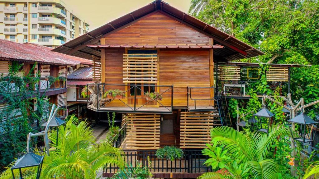 A charming wooden house is showcased, featuring slatted shutters and balconies adorned with plants. Surrounded by lush greenery and pathways, the structure exhibits a blend of natural elements and architectural design, complemented by soft lighting from nearby lamps.