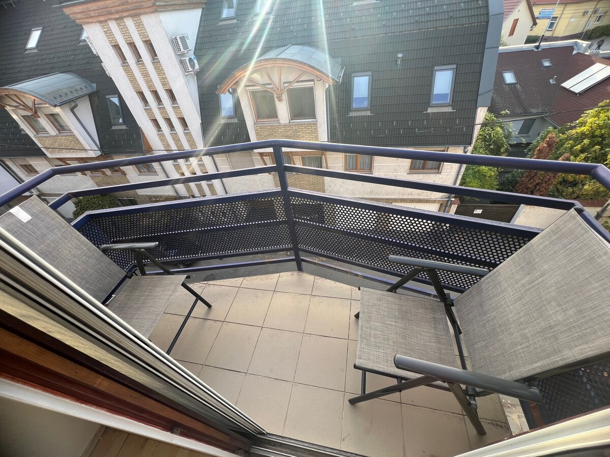 A private balcony is presented with two minimalistic chairs and a protective railing. A view of neighboring buildings can be seen beyond the balcony, illuminated by sunlight, creating a relaxed outdoor space.