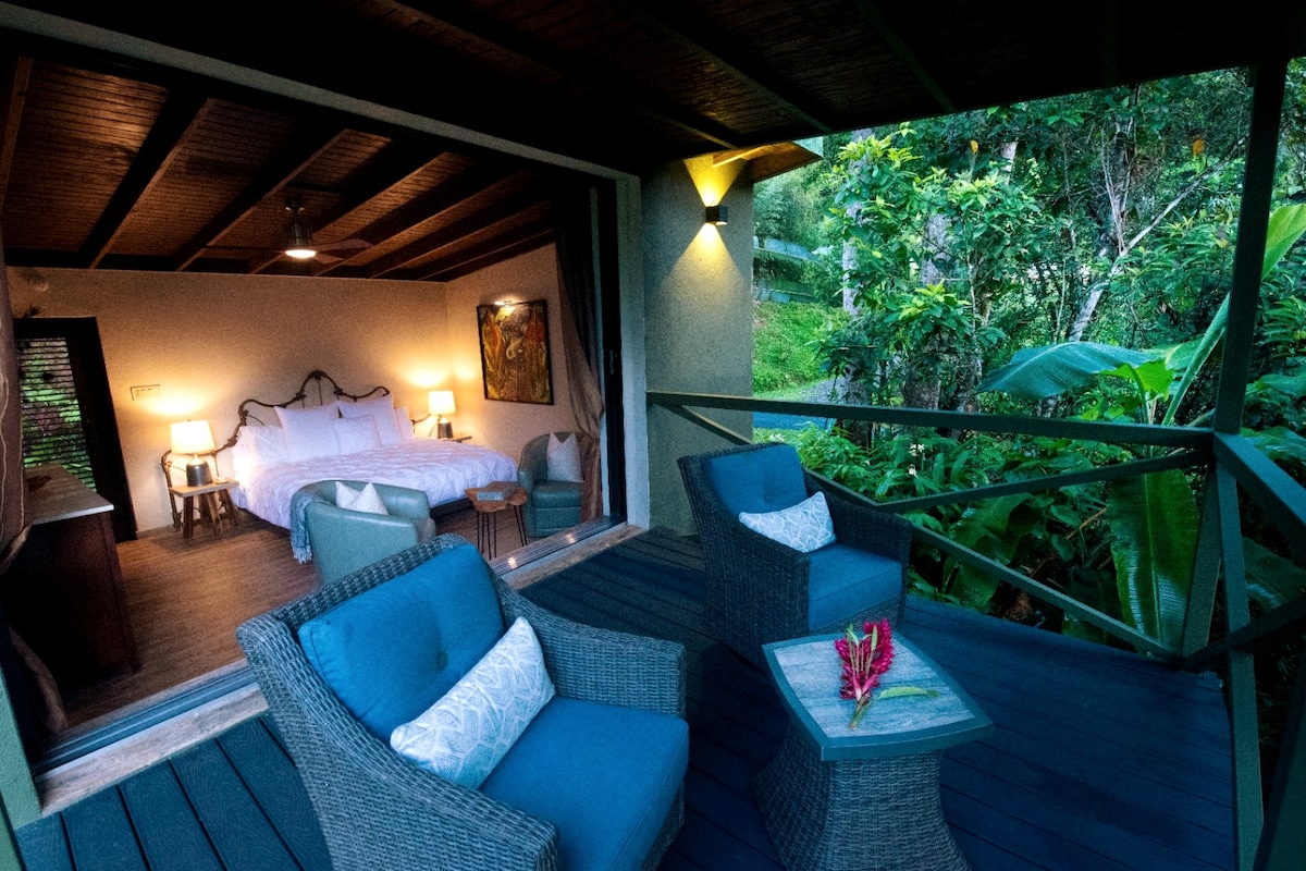 A private terrace is furnished with two comfortable armchairs and a small table, adorned with a vibrant flower. In the background, a spacious bedroom is visible, featuring a light-colored bed and warm lighting, surrounded by lush greenery.