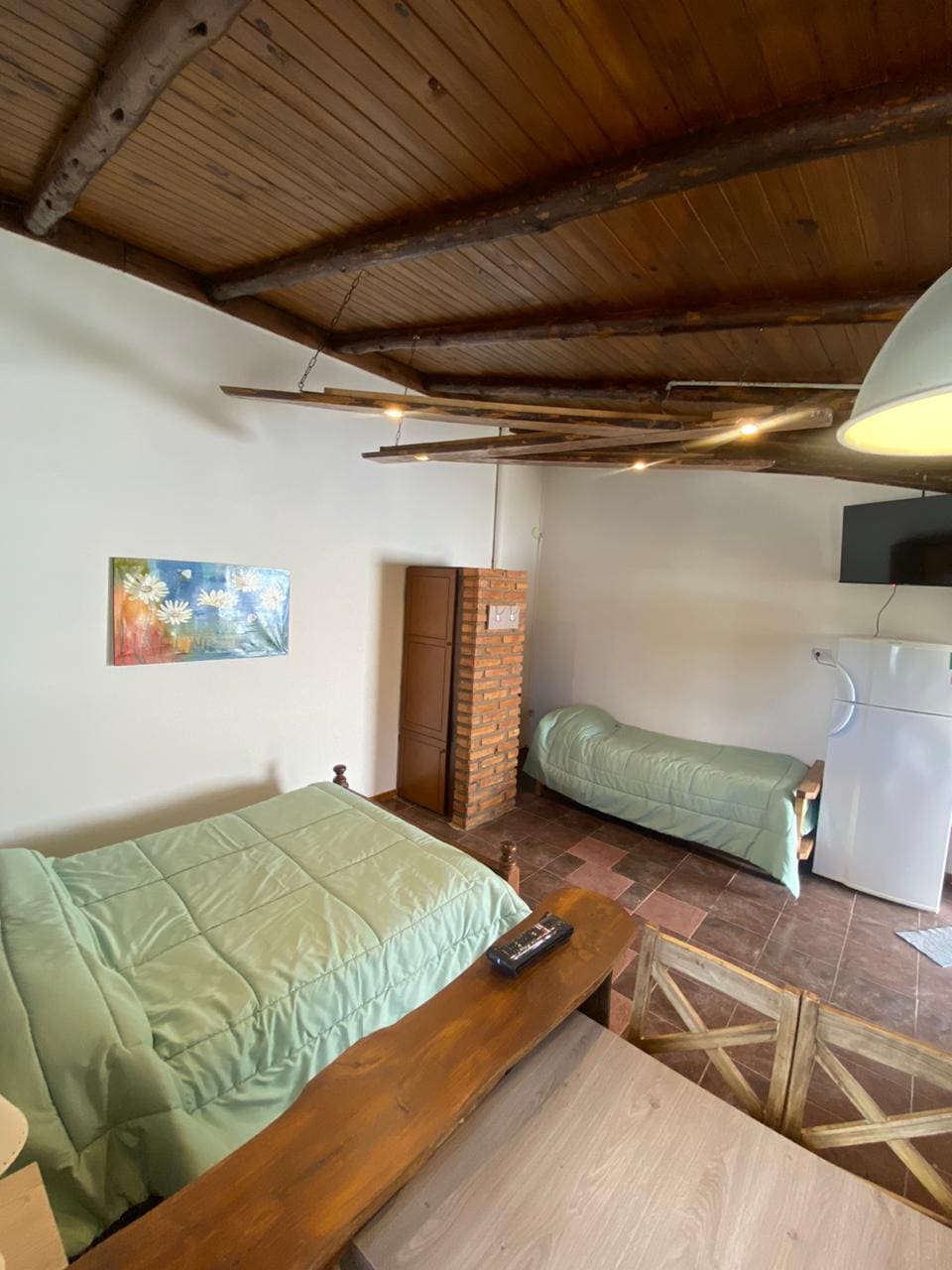 The room includes two beds with light green bedding, positioned against a wooden wall. A small wooden table is situated in the foreground. A piece of artwork is displayed on one wall, and a refrigerator is visible in the background.