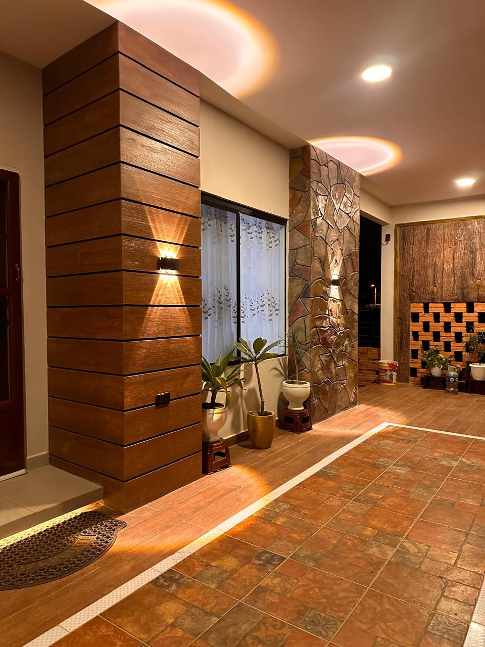 The entryway features a combination of natural wood and stone elements, highlighted by warm lighting. Potted plants are arranged along the sides, adding a touch of greenery. The floor showcases an intricate tile pattern, leading towards a softly lit area in the background.