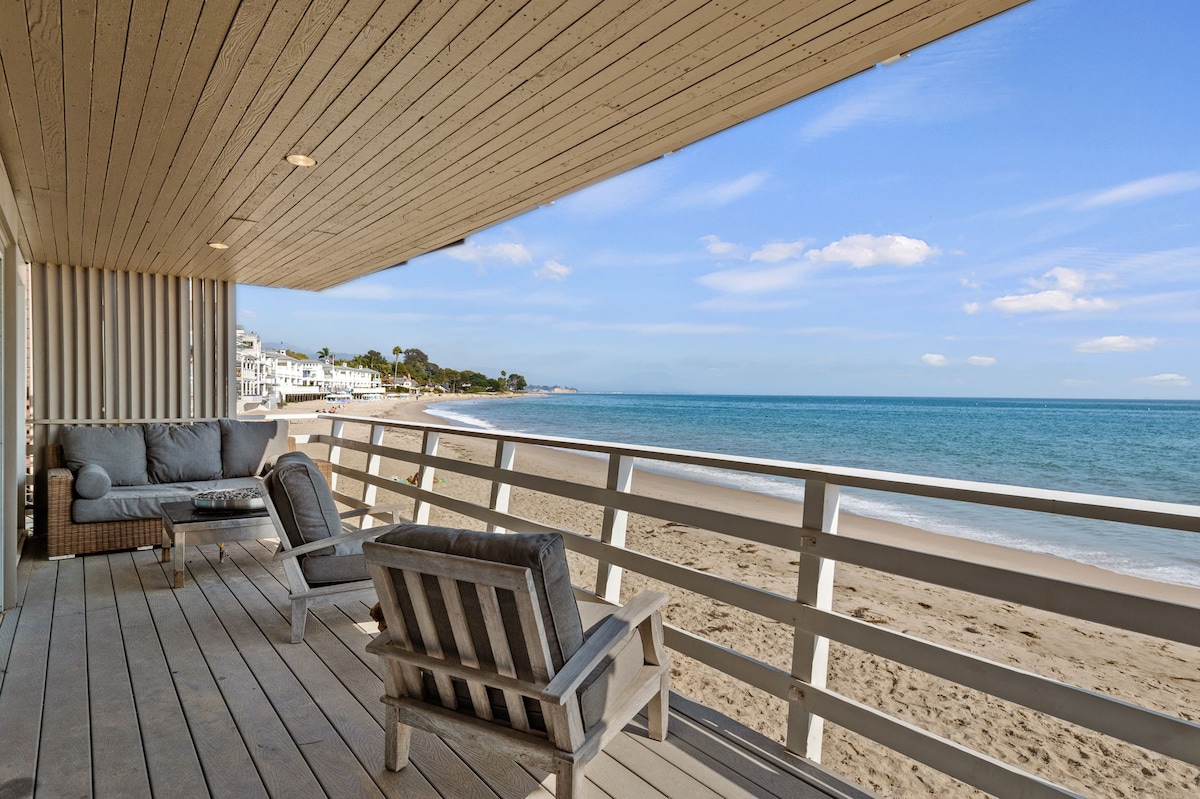 A private deck is showcased, featuring comfortable seating and a coffee table, all positioned to offer unobstructed views of Miramar Beach. Soft sand and calm ocean waves are visible, enhancing the tranquil setting. The structure is highlighted by the stylish wooden ceiling and railing.