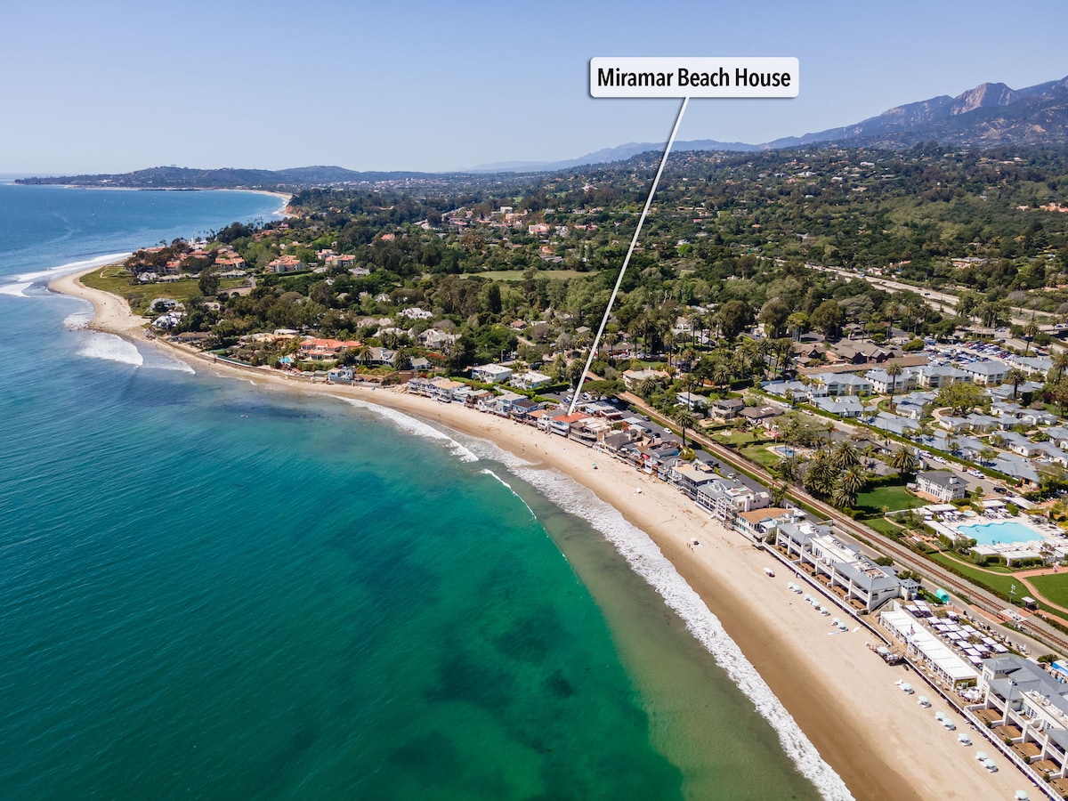 An aerial view captures the coastline of Miramar Beach, showcasing the sandy shore meeting the turquoise waters. The beach house is positioned near the water's edge, with nearby roads and lush greenery visible in the background, providing a sense of the location's natural beauty.
