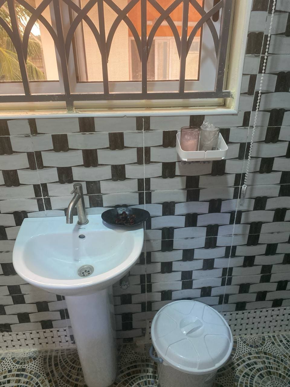 A compact bathroom setting is showcased with a modern sink mounted on a pedestal. The sleek faucet complements the black and white patterned tiles. A toilet is positioned nearby, while a shelf holds toiletries. Natural light filters through the patterned window, enhancing the space.