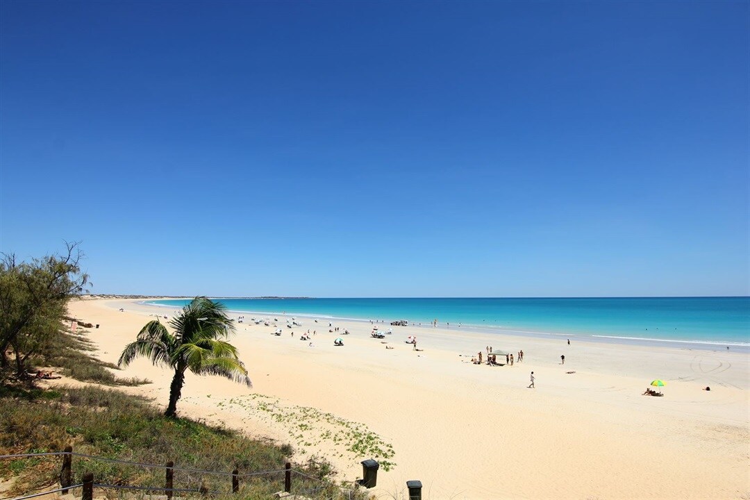 A wide sandy beach is captured under a clear blue sky, with gentle waves lapping at the shore. Sunbathers and umbrellas dot the landscape. Lush palm trees are present along the beach, adding a touch of greenery to the coastal scene.
