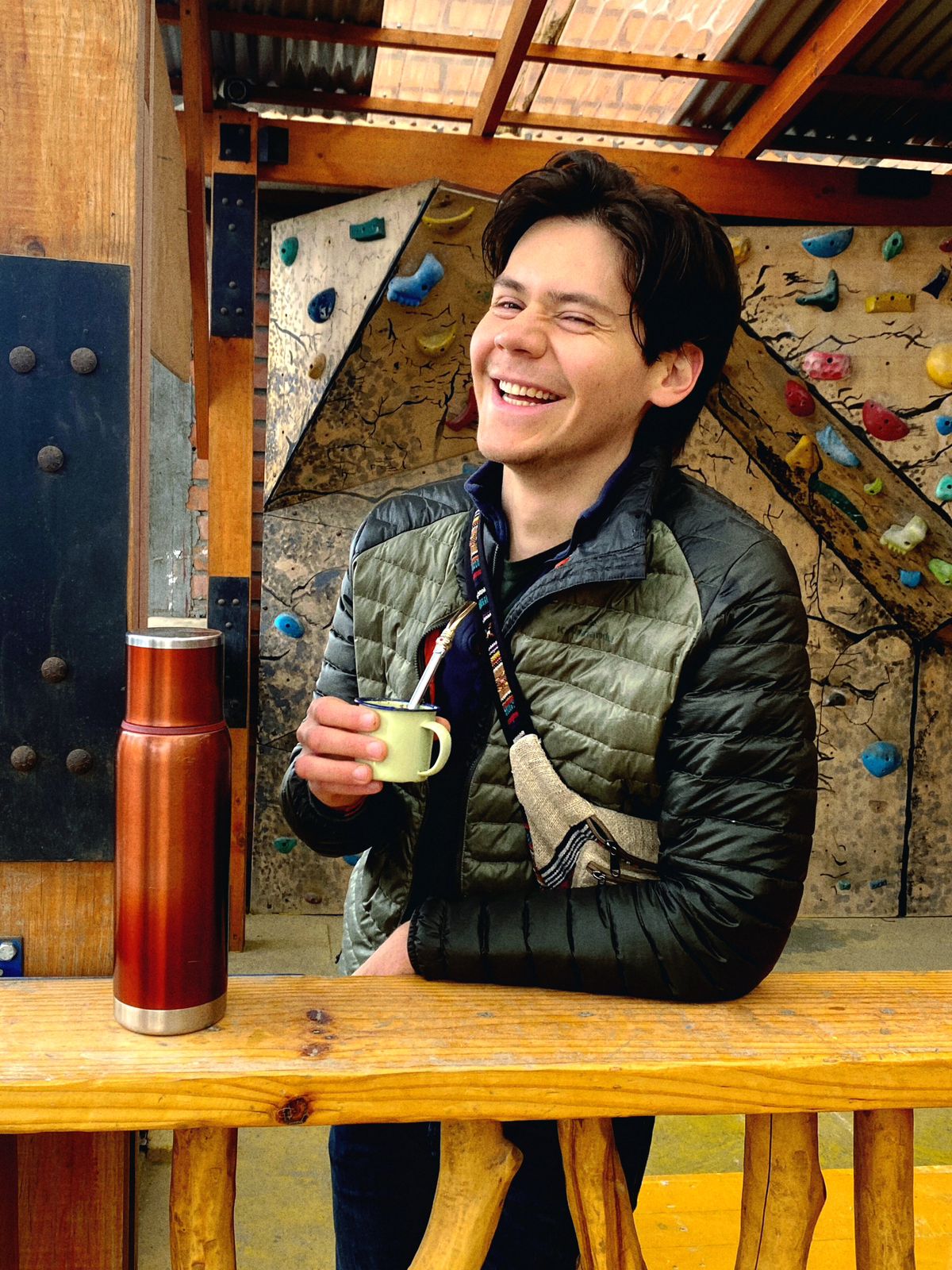 A person stands near a wooden counter, holding a small cup. A stainless steel thermos is positioned next to them, with colorful climbing holds visible on a nearby wall. The setting features natural materials and offers a casual atmosphere.