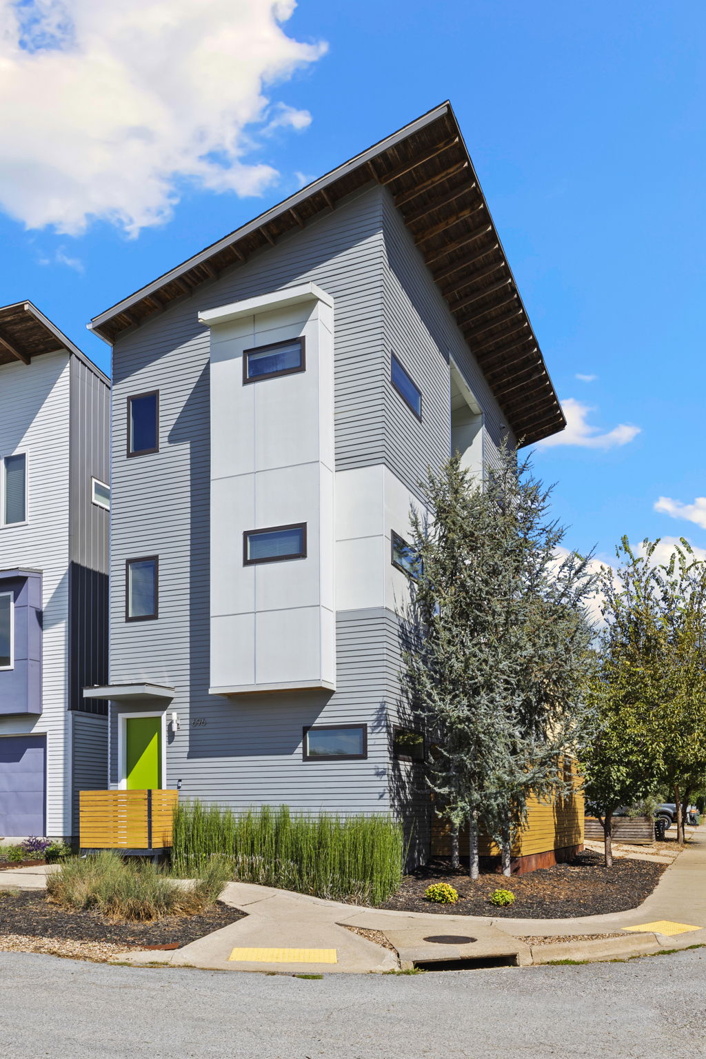 The exterior of the house displays a modern architectural design with multiple levels and large windows. A vibrant green door serves as a focal point, while surrounding landscaping features low-maintenance shrubs and small trees. The crisp blue sky adds a bright backdrop.