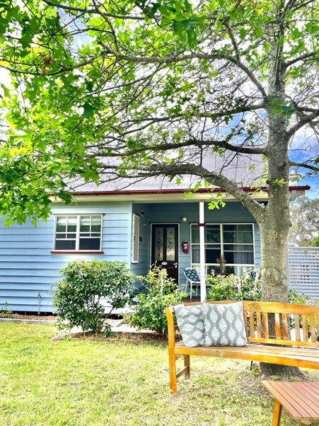 The cottage exterior is framed by a mature tree, offering shade to a wooden bench with a patterned cushion. The light blue siding and large windows enhance its charm, while a small outdoor seating area is visible, surrounded by well-maintained greenery.