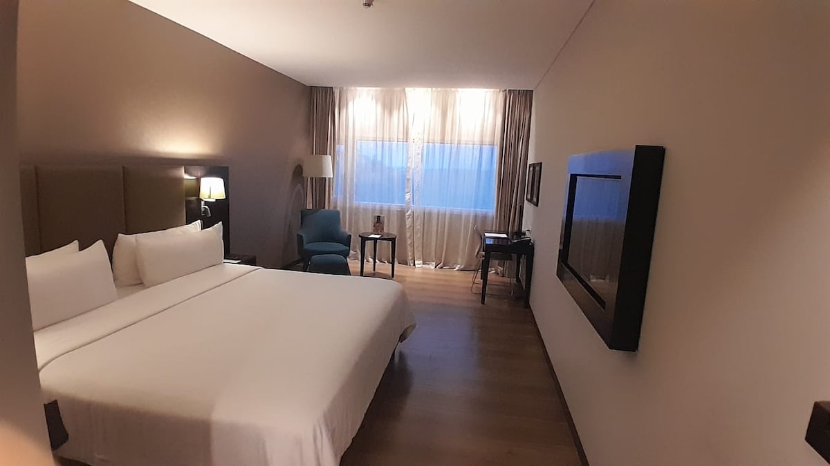 A spacious hotel room features a large bed dressed in crisp white linens. A comfortable armchair is positioned beside a window with sheer curtains, allowing natural light to filter in. A wall-mounted television and a small desk complete the contemporary decor.