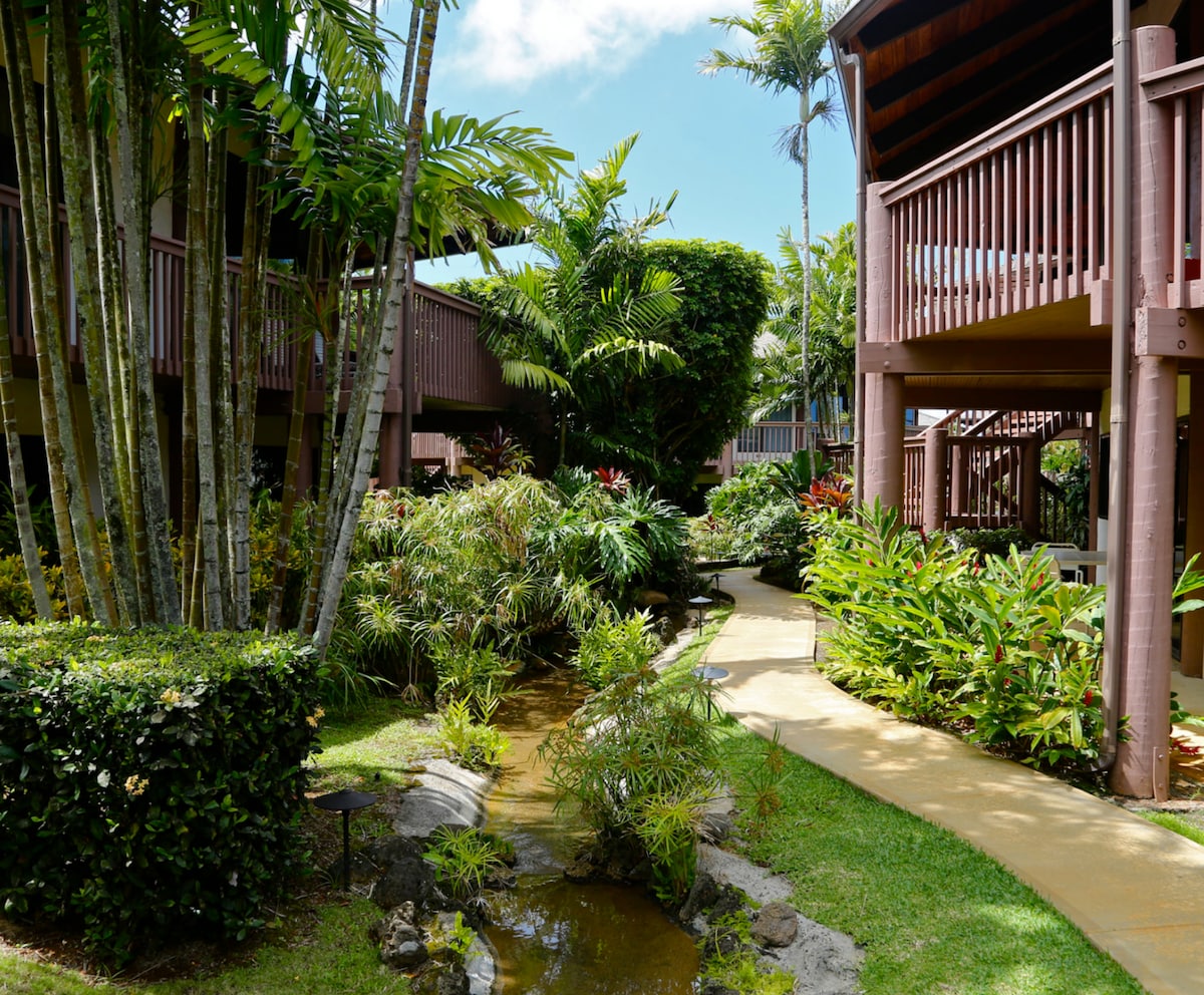 A tropical garden pathway is framed by lush greenery, featuring tall trees and vibrant plants. The pathway curves through the garden, leading to wooden balconies and charming buildings that nestle among the foliage, reflecting a tranquil atmosphere.