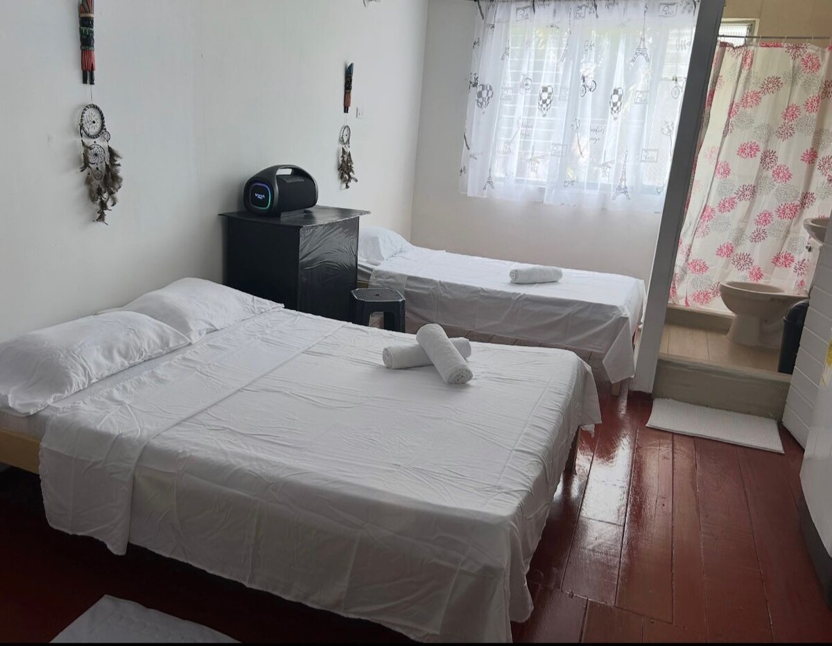 A bright sleeping area features two spacious beds covered with white linens. A small black cabinet is positioned beside a window adorned with sheer curtains that allow natural light to enter. A bathroom area is visible, separated by a decorative shower curtain.
