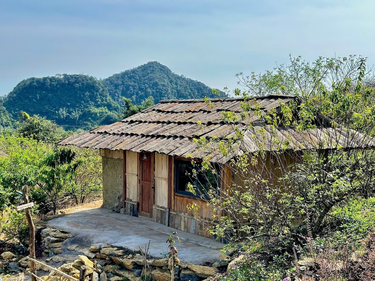 A traditional Hmong homestay is surrounded by greenery, featuring a rustic wooden exterior with a thatched roof. The structure is set against a serene backdrop of rolling mountains, creating a very peaceful ambiance. A stone pathway leads up to the entrance.