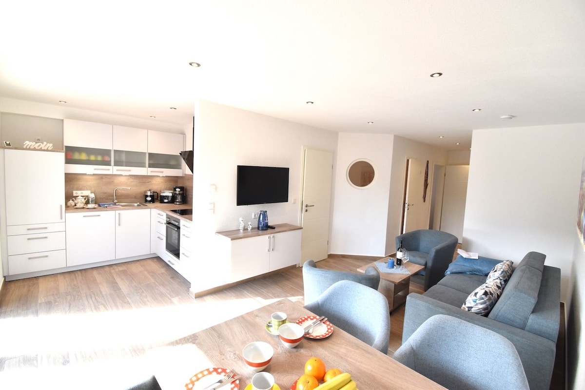 The open-plan living area features a modern kitchenette with sleek cabinetry and a dining table set for breakfast. A comfortable seating arrangement with light blue sofas offers a casual gathering space. Natural light fills the room through well-placed lighting fixtures.