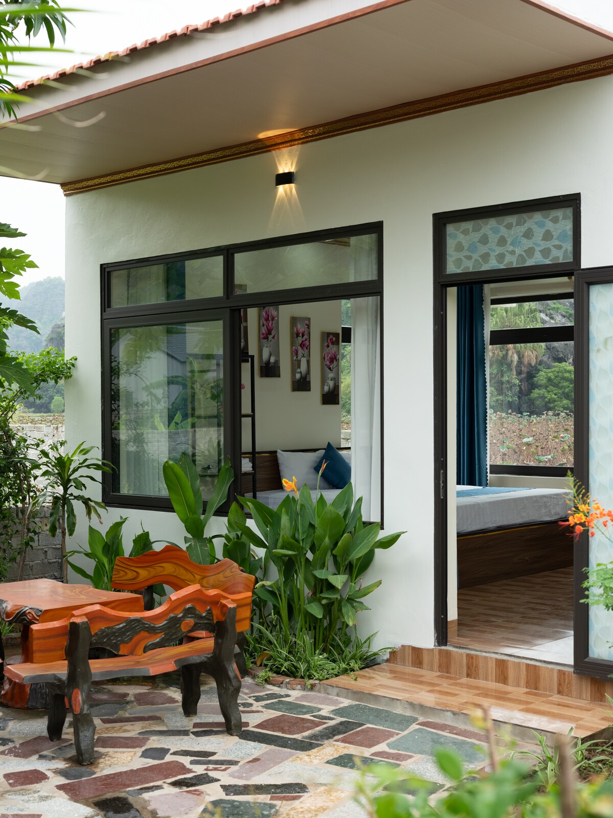An inviting outdoor seating area is positioned next to a lush garden, featuring a wooden table and benches. Large windows frame the entrance, allowing natural light to brighten the interior space, which includes views of the mountains and greenery.