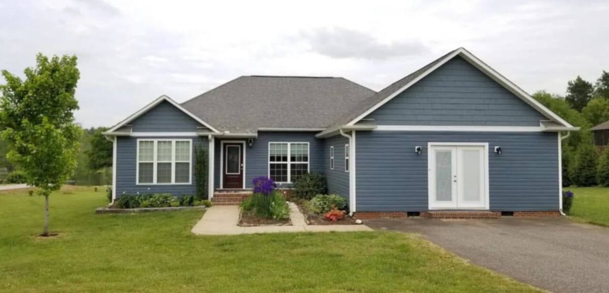A single-story blue house with a gray shingle roof is surrounded by well-maintained lawns and gardens. Bright flowers and shrubs enhance the entryway, complemented by large windows that provide natural light. A concrete walkway leads to a welcoming front porch area.