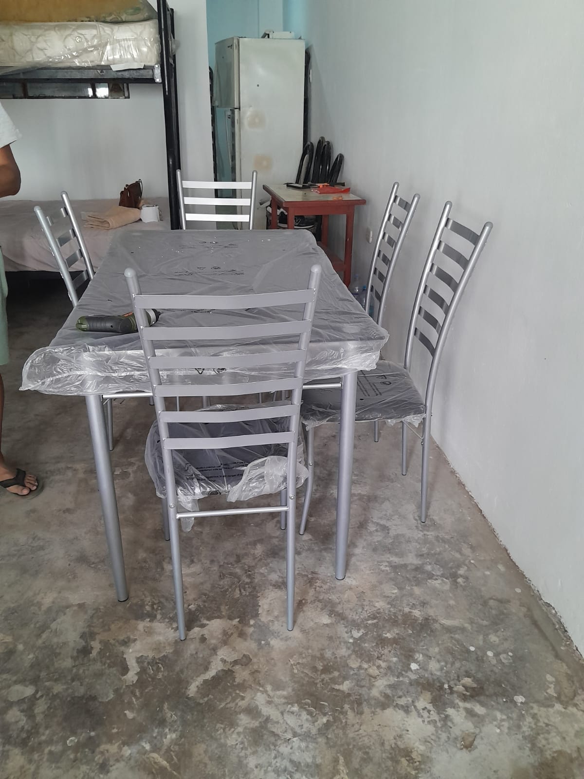 A simple dining area features a rectangular table surrounded by five silver chairs. The table is covered with a protective layer. A fridge and kitchen utensils can be seen in the background, against a plain wall.