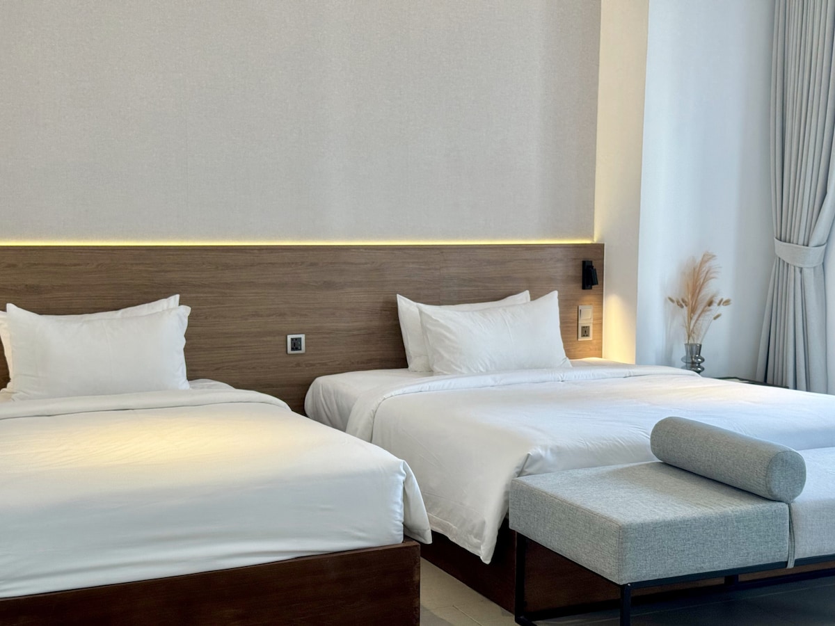 A well-lit room features a spacious queen bed and a single bed, both dressed in crisp white linens. A minimalist bench sits at the foot of the beds, while soft curtains frame the window, allowing natural light to enhance the neutral tones of the decor.