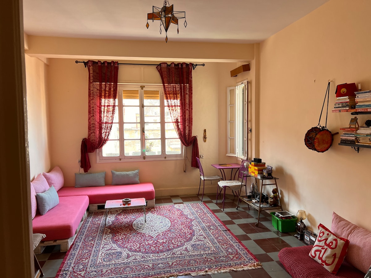 A living room is presented with a vibrant red sofa and decorative cushions. A small coffee table sits on a patterned area rug, while a decorative bookshelf and side table add functional elements. Natural light filters through multiple windows, complemented by flowing curtains.