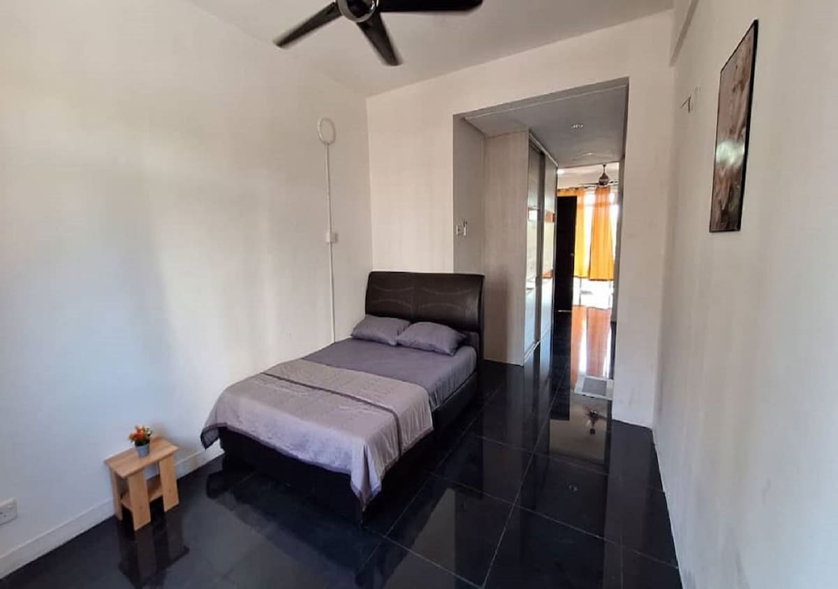 A simple sleeping area features a spacious bed with a dark headboard, dressed in a gray blanket. A small wooden side table with a plant adds a touch of decor. The room is illuminated by natural light, reflecting off the polished black tile floor.