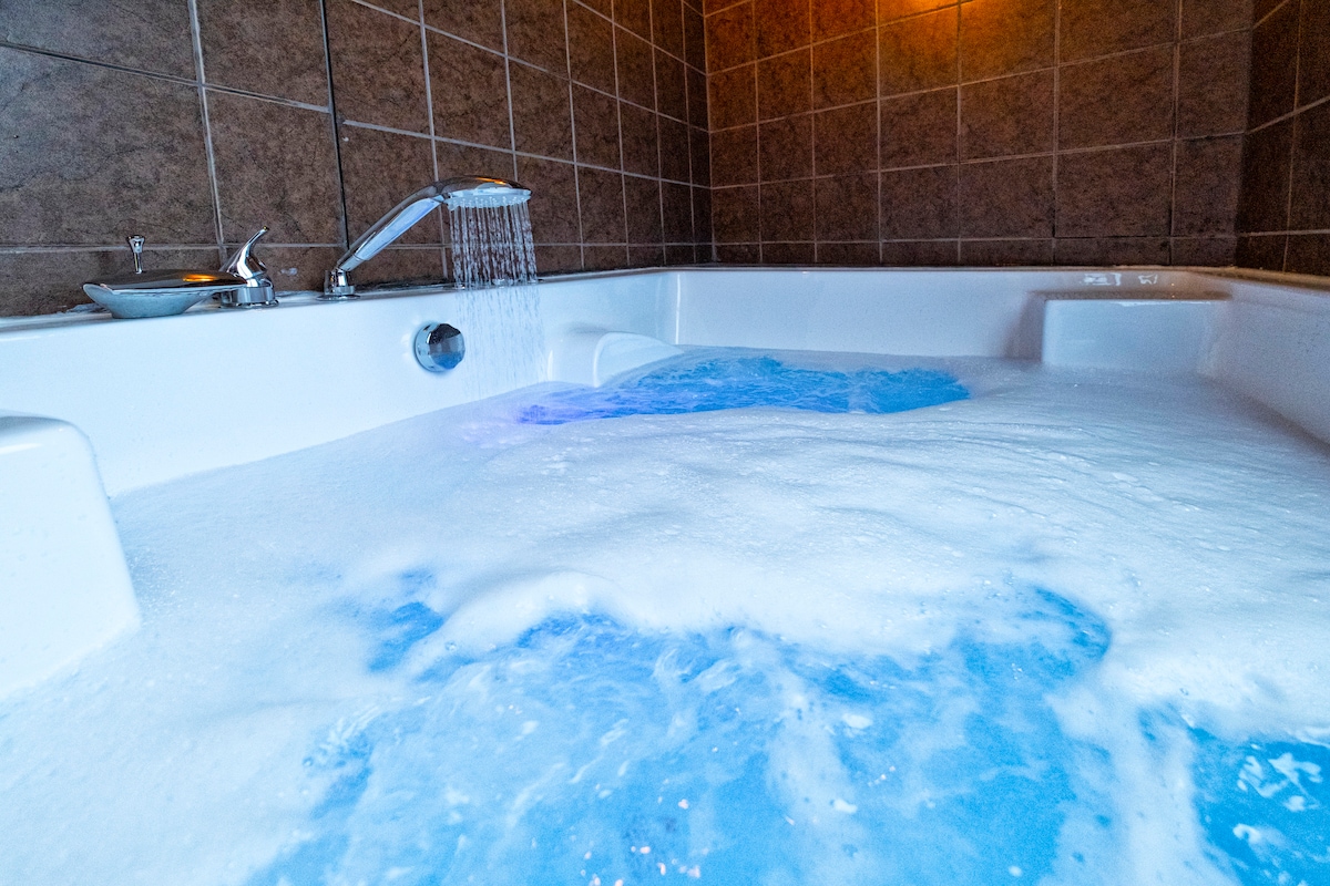 A spacious hot tub is filled with bubbles and illuminated by soft blue lighting, creating a serene atmosphere. The tiled walls and subtle overhead lighting enhance the overall design, contributing to a relaxing environment.