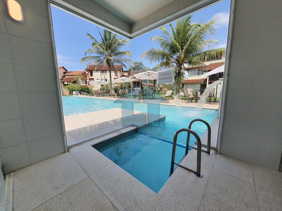 A spacious indoor area features a large window that frames the pool outside. The pool area is surrounded by palm trees and lounging chairs, with a serene atmosphere highlighted by blue skies. Water gently cascades from a small feature near the entry.